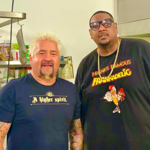 Guy Fieri and a man in a Frank's Famous Chicken & Waffles shirt pose in a kitchen.