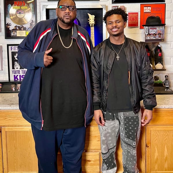 Two men, one older and one younger, stand smiling in front of music memorabilia at Frank's Famous.
