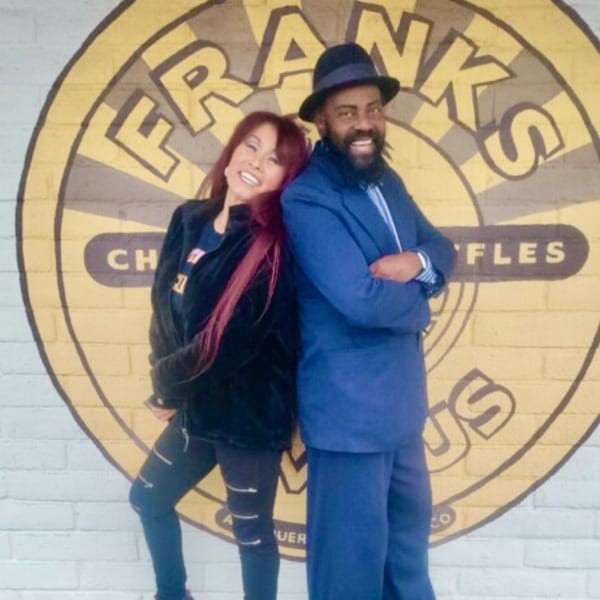 Two smiling people, a woman and a man, pose in front of the Frank's Famous Chicken & Waffles logo.