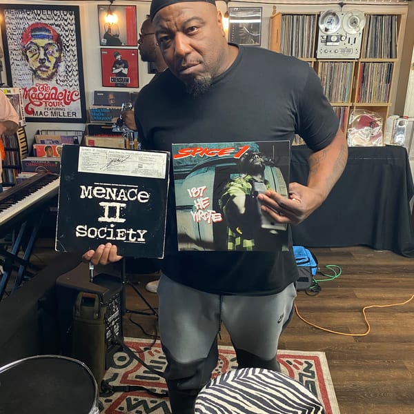 Man in a black shirt holding two vinyl records, "Menace II Society" and "Spice 1," in a room with music memorabilia.