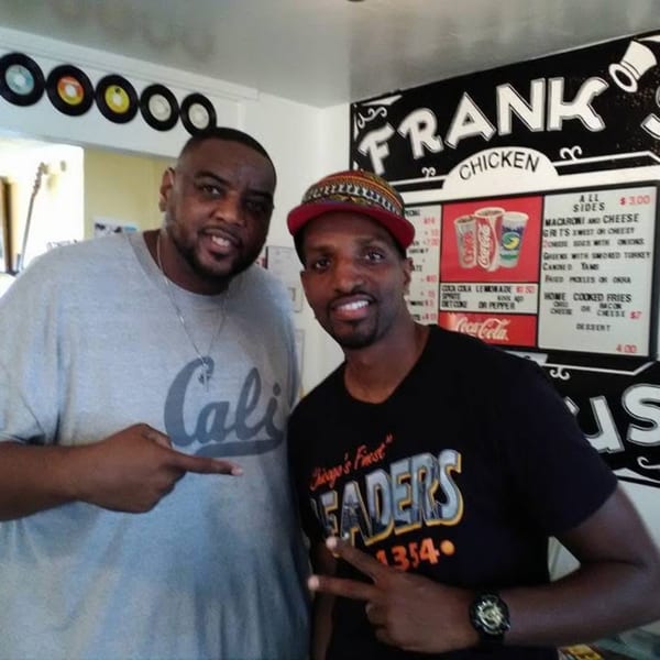 Two men smiling, one pointing, in front of a Frank's Famous Chicken & Waffles menu.