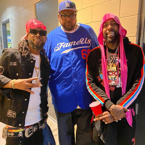 Three men in casual attire, one in a blue "Famous" jersey, posing for a photo.