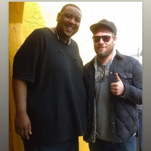 Two men, one smiling broadly, the other giving a thumbs-up, stand against a yellow wall.