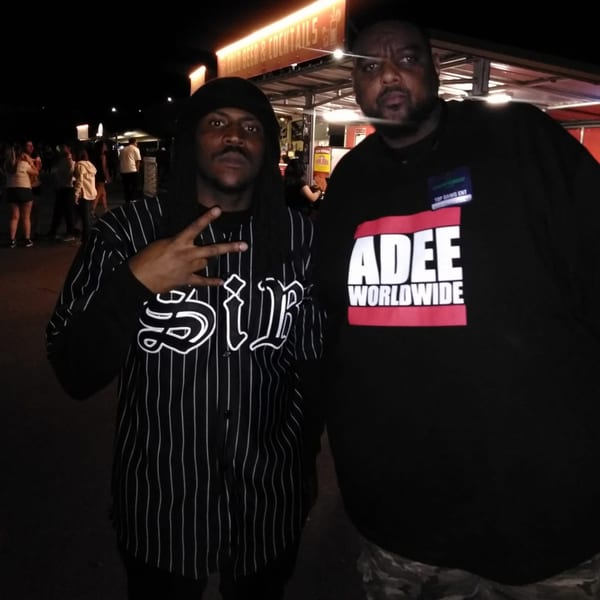 Two men pose for a photo at night, one in a striped shirt, the other in a black tee.