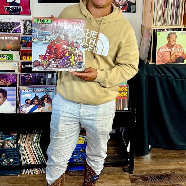 A person in a North Face hoodie holds a Bill Cosby record album in a room filled with vinyl.