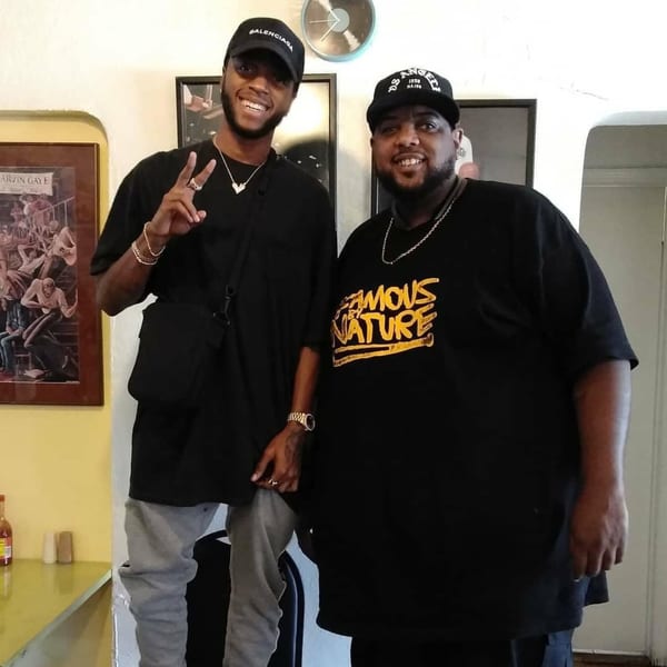 Two men, one wearing a Balenciaga hat, smiling and posing inside Frank's Famous Chicken & Waffles.