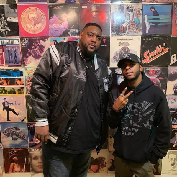 Two men posing in front of a wall covered with vintage vinyl record albums.