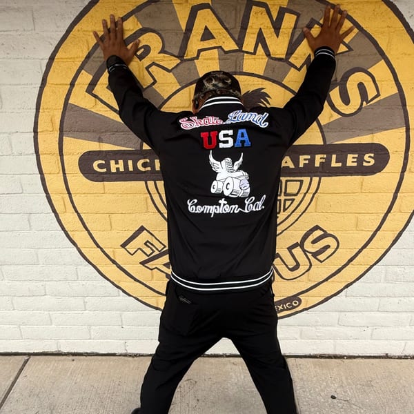Person in black jacket with "Skate Land USA Compton Cal" on back, arms raised against Frank's Famous mural.
