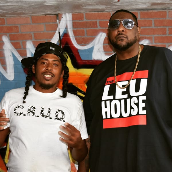 Two men, one in a white shirt and the other in a black shirt, stand before a brick wall.