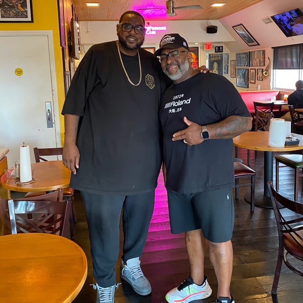 Two men posing together inside Frank's Famous Chicken & Waffles restaurant, with neon signs and tables.