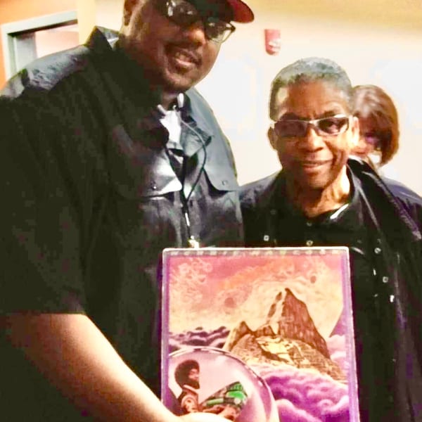 Two men, one wearing a cap and sunglasses, holding a colorful book with a mountain on its cover.