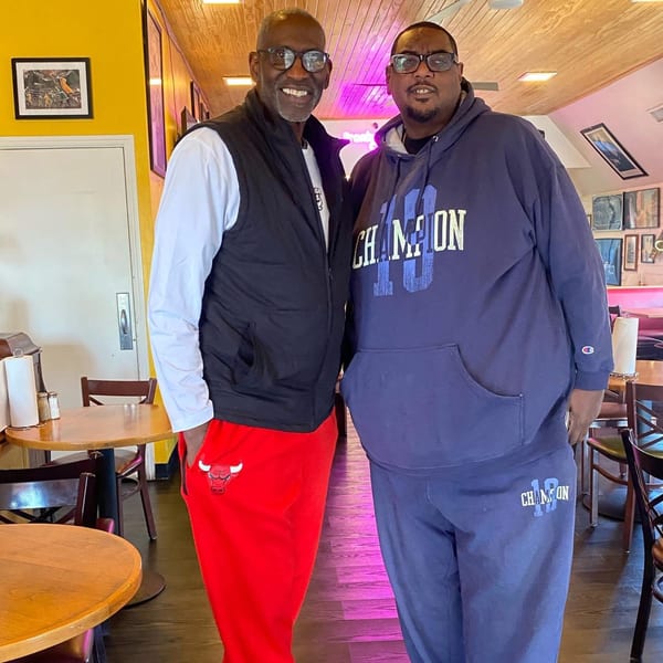 Two men smiling, standing in a brightly lit Frank's Famous Chicken & Waffles dining room.