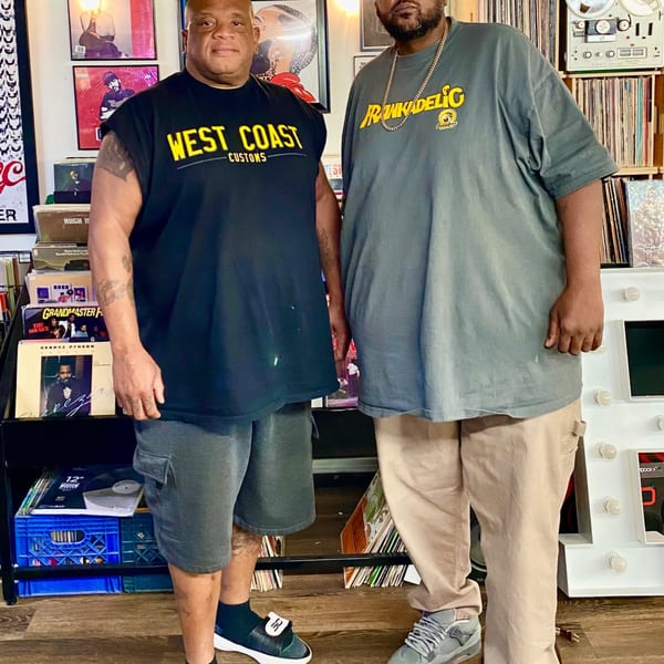 Two men stand in a room filled with vinyl records, one wearing a "West Coast Customs" shirt.