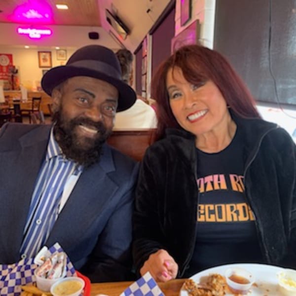 Two smiling customers, a man in a fedora and a woman, enjoying chicken and waffles at Frank's Famous.