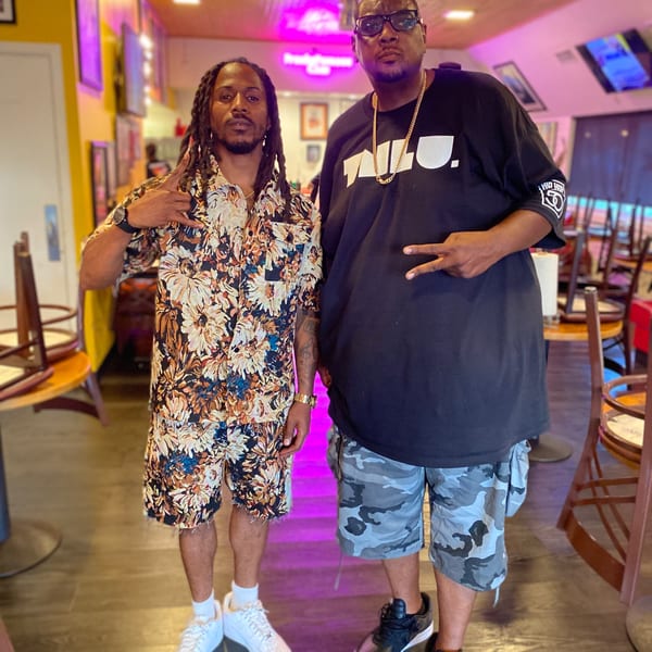Two men posing in a restaurant with neon pink lighting and wooden chairs.