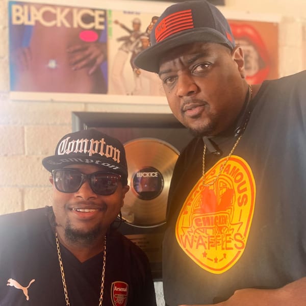 Two men, one wearing a Frank's Famous Chicken & Waffles shirt, pose in front of records.