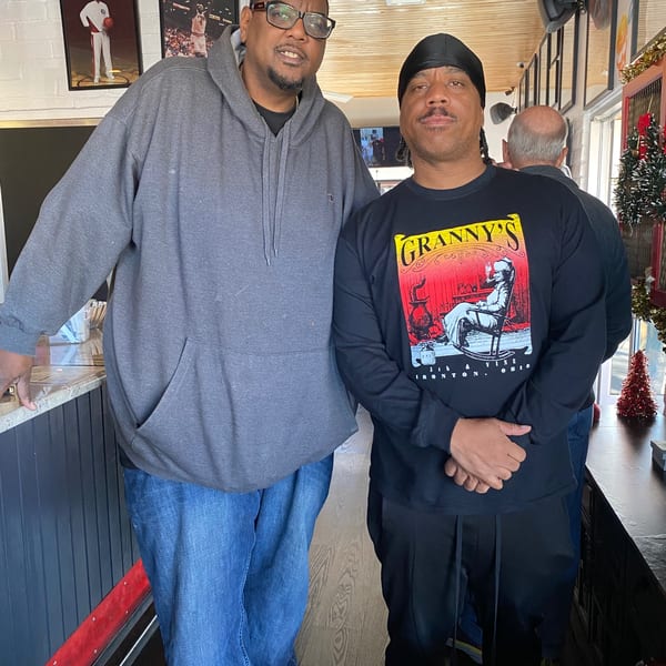 Two men posing inside Frank's Famous Chicken & Waffles, with framed sports photos on the wall.