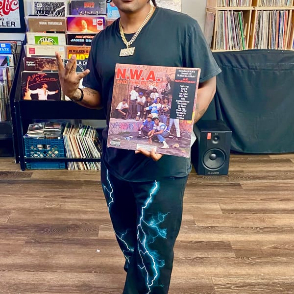 A person in a black shirt and lightning-patterned pants holds an N.W.A. record album in a record store.
