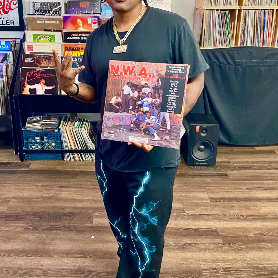A person in a black shirt and lightning-patterned pants holds an N.W.A. record album in a record store.