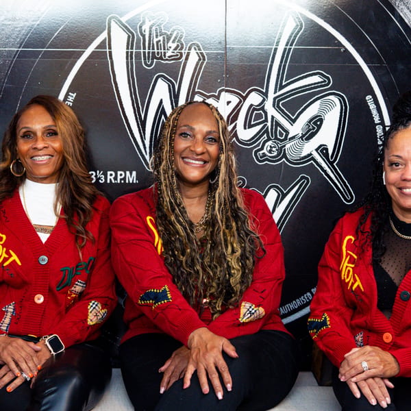 Three smiling women in red cardigans with patches, sitting in front of a large record graphic.