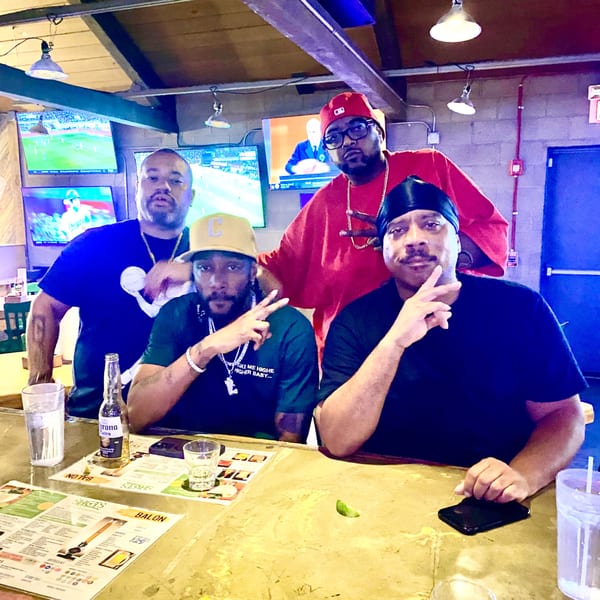 Four men pose for a photo at a bar, with drinks and menus on the counter.