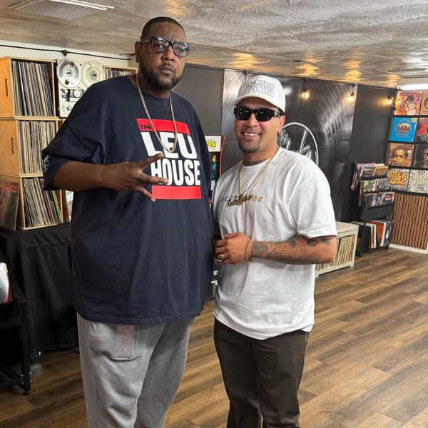 Two men, one in a black t-shirt and the other in a white t-shirt, stand in a room with records.