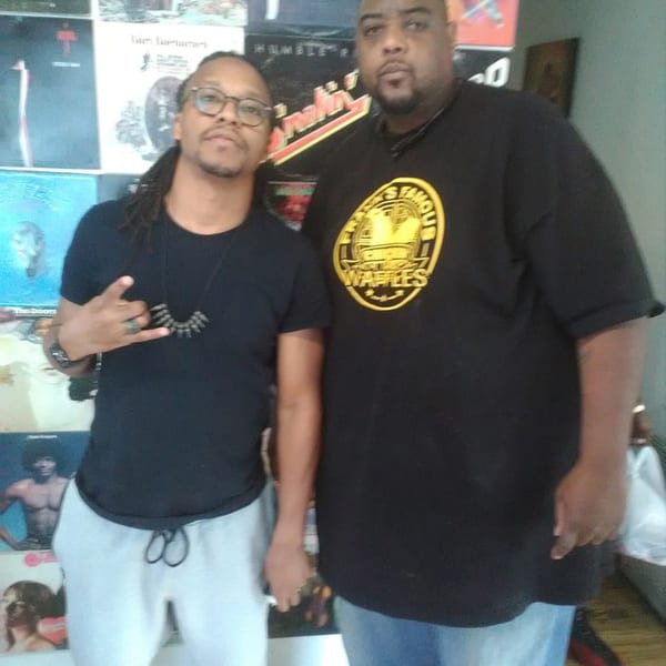 Two men stand together, one wearing a Frank's Famous Chicken & Waffles t-shirt.