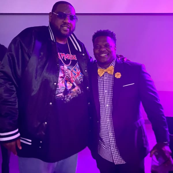 Two men posing for a photo under purple lighting, one in a varsity jacket, the other in a suit.