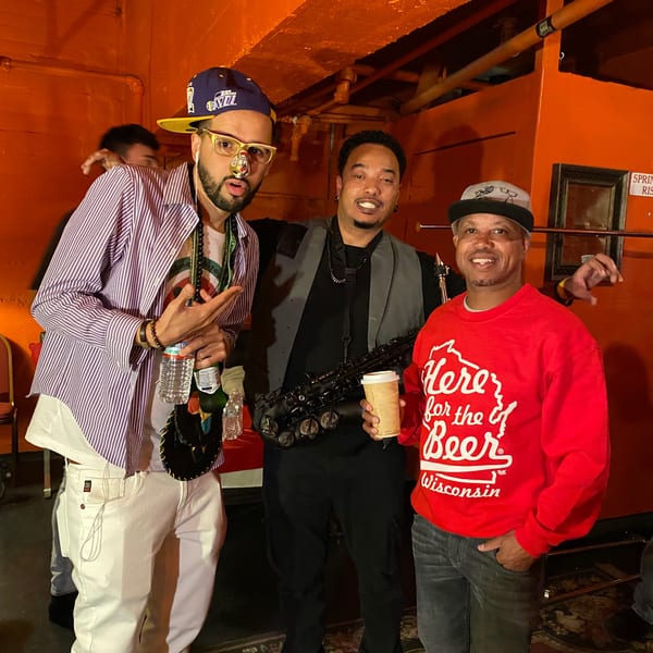 Three men posing for a photo, one holding a saxophone, against an orange wall.