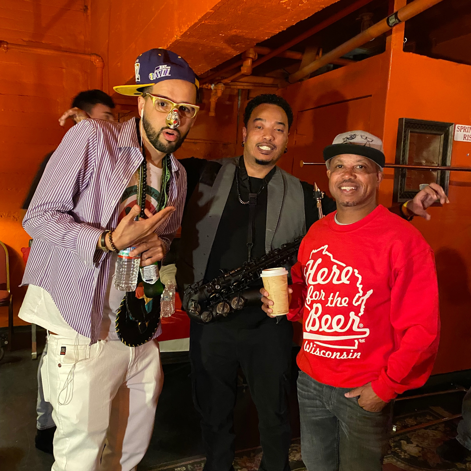 Three men posing for a photo, one holding a saxophone, against an orange wall.